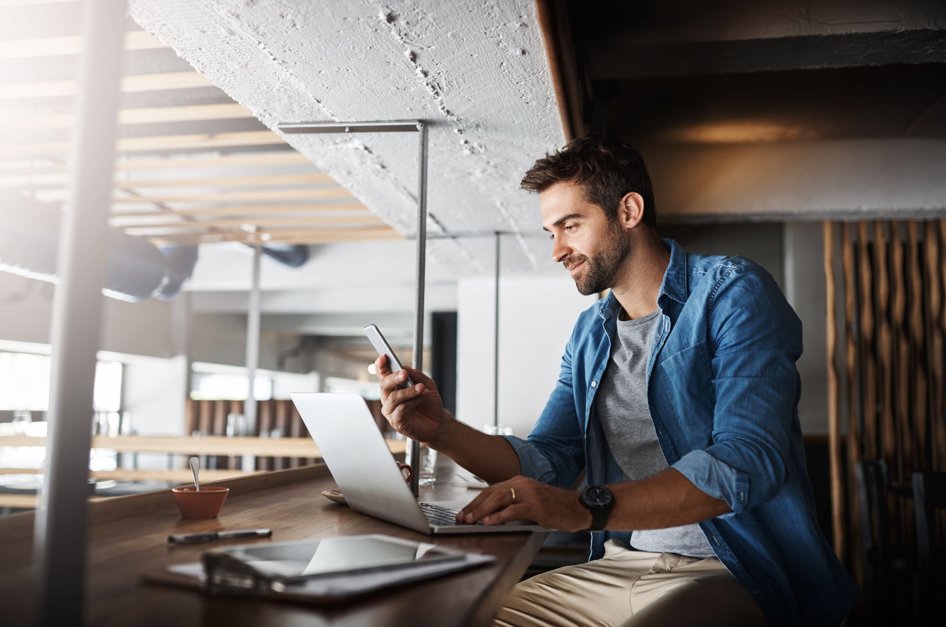 A man is sitting at a table using a laptop and a cell phone.