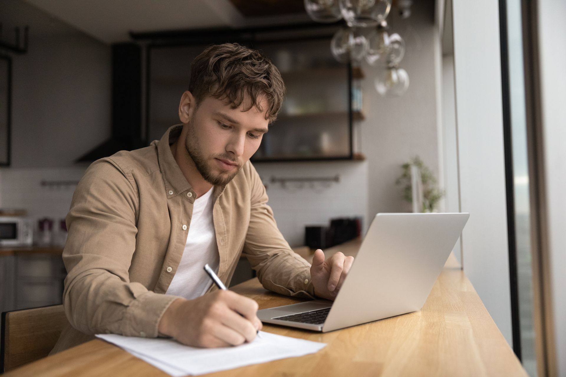 A man is sitting at a table with a laptop and writing on a piece of paper.