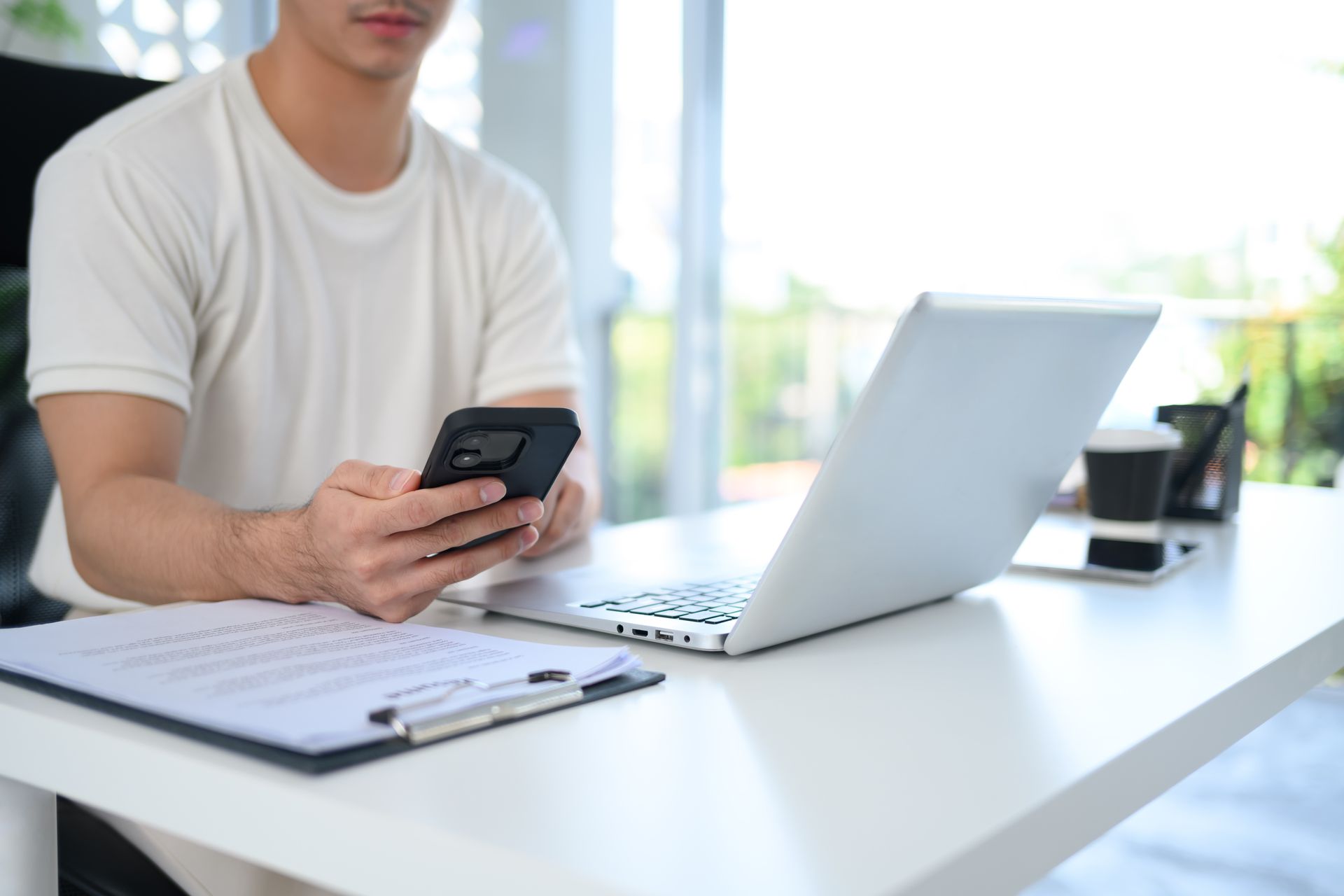 A man is sitting at a desk using a laptop and a cell phone.