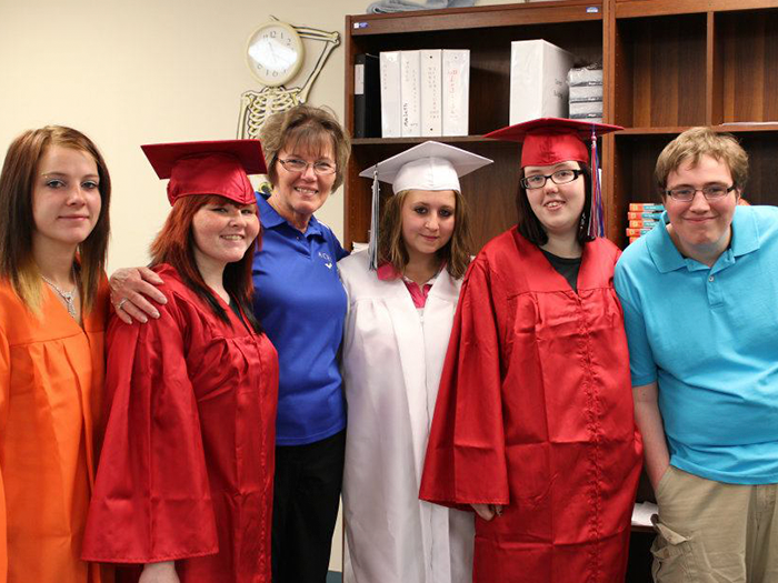 Group of graduates in caps and gowns, posing with an adult, in a room with shelves and a skeleton model.
