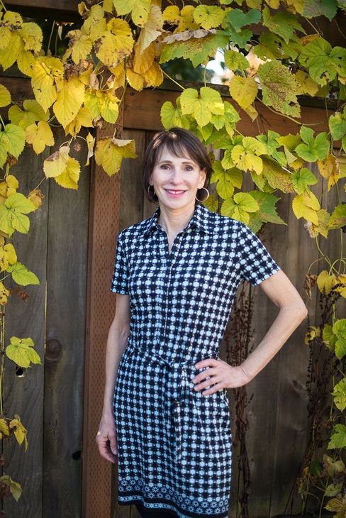 Woman in checked dress stands near wooden fence with fall foliage.