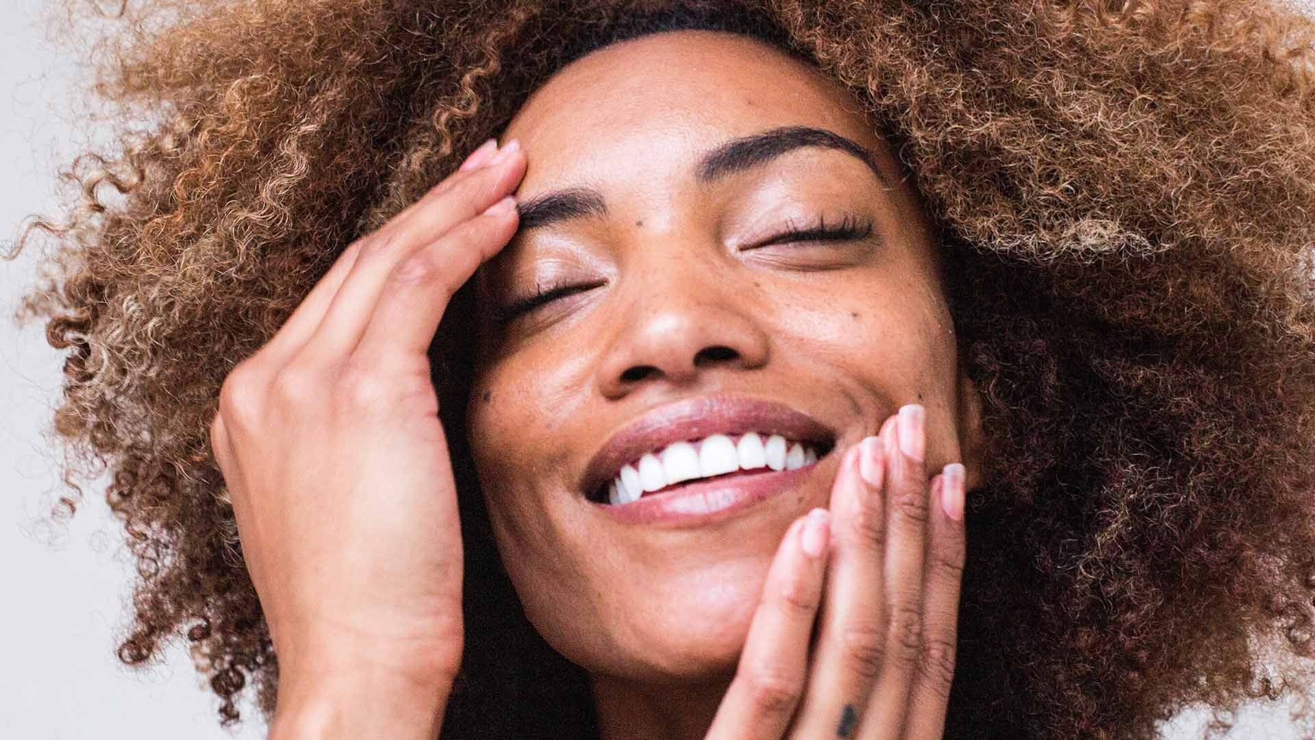 Woman with curly hair smiles, eyes closed, touching face.