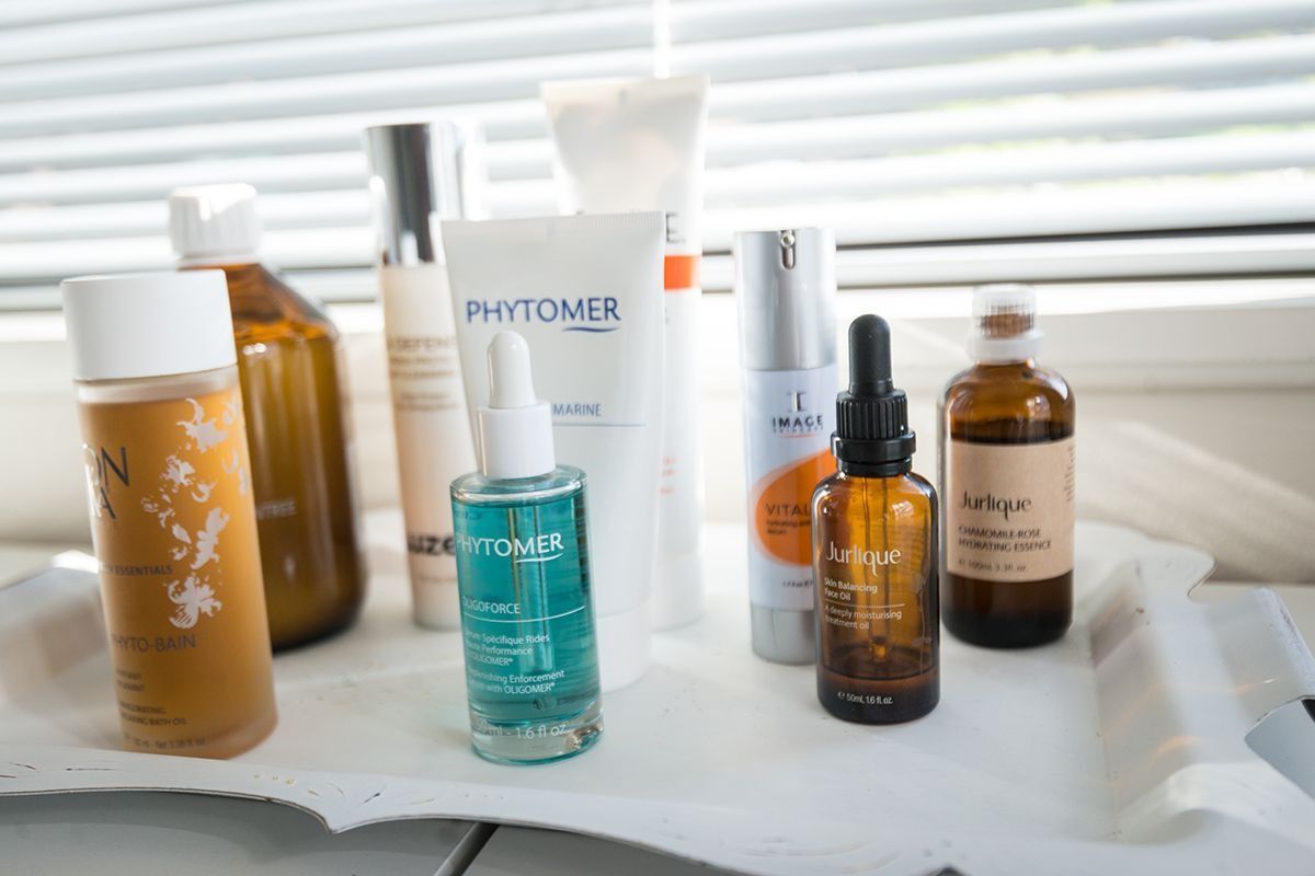 Skincare products: bottles, serums, and tubes, on a white tray by a window with blinds.