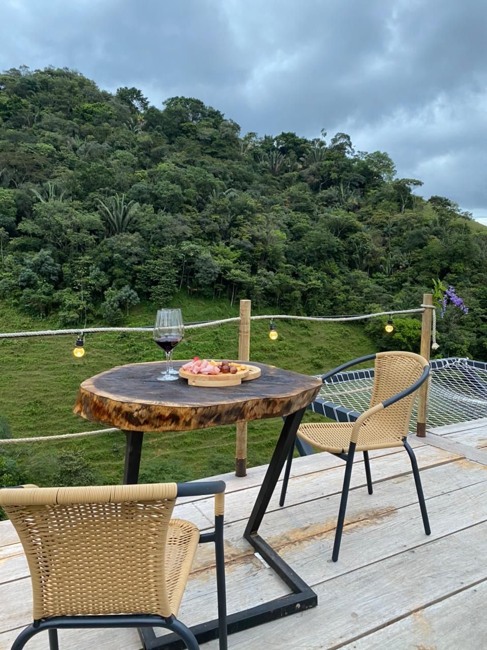 Mesa de madera con comida y vino, dos sillas, con vistas a una ladera verde.