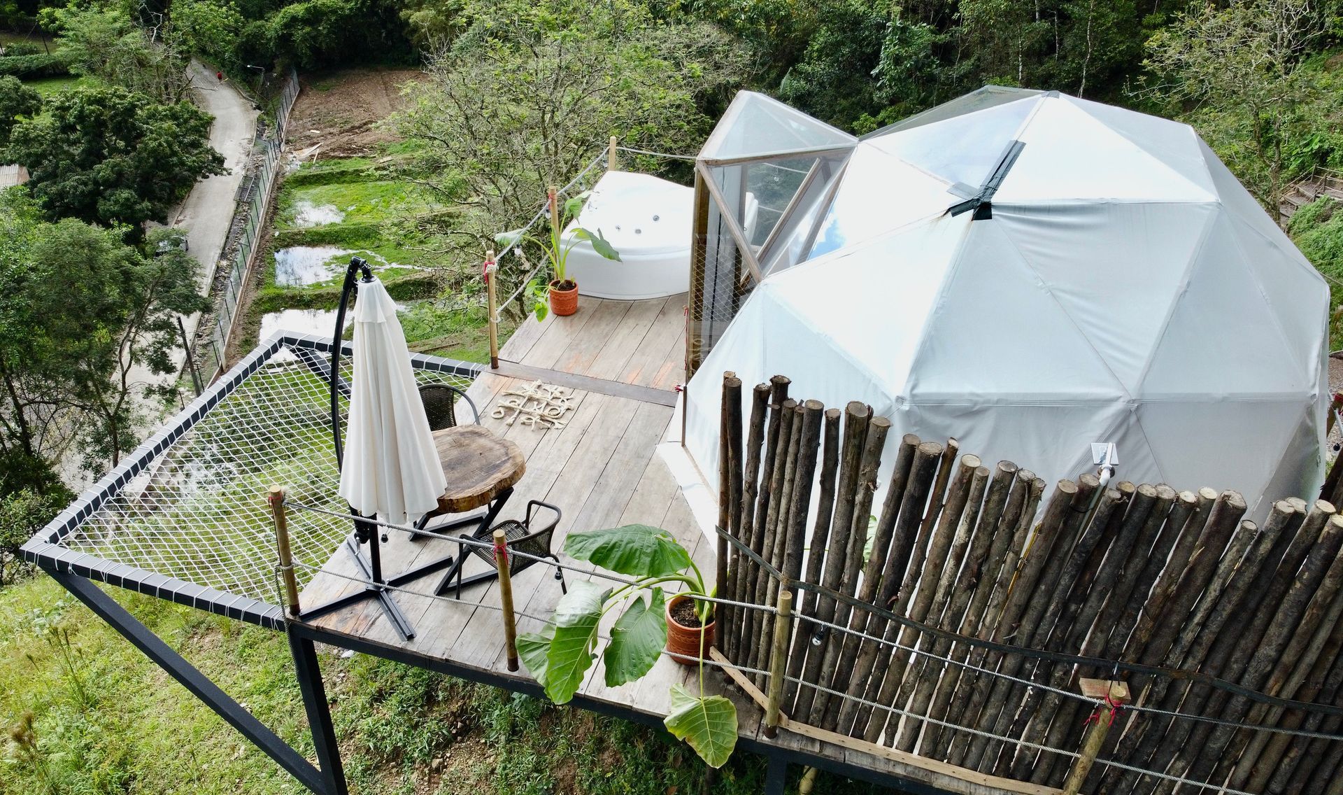 Una estructura glamping con cúpula geodésica con plataforma de madera, sombrilla y cerca de bambú en un entorno natural.
