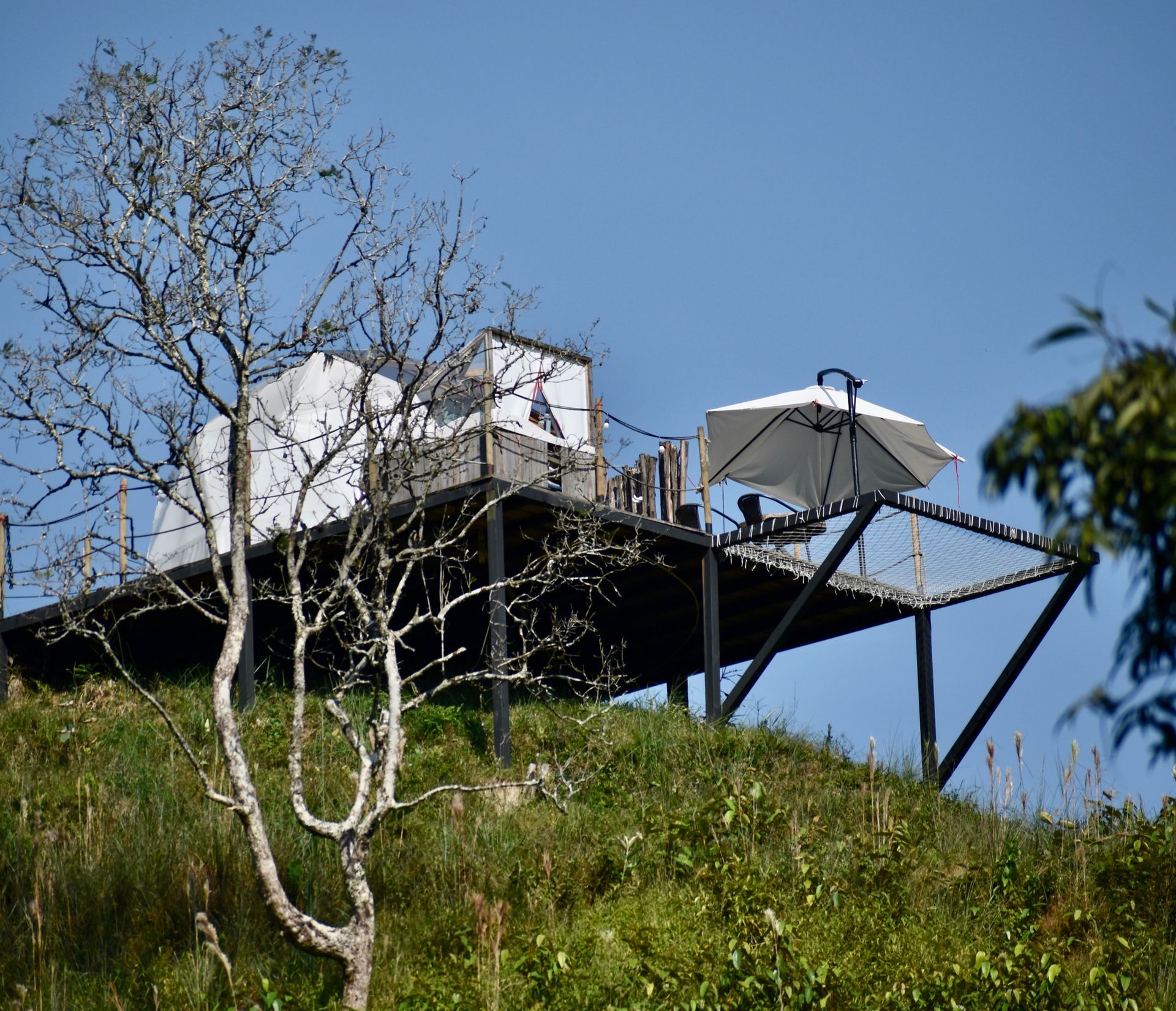 Cabaña elevada en una ladera con terraza, sombrilla y un árbol en primer plano contra un cielo azul.