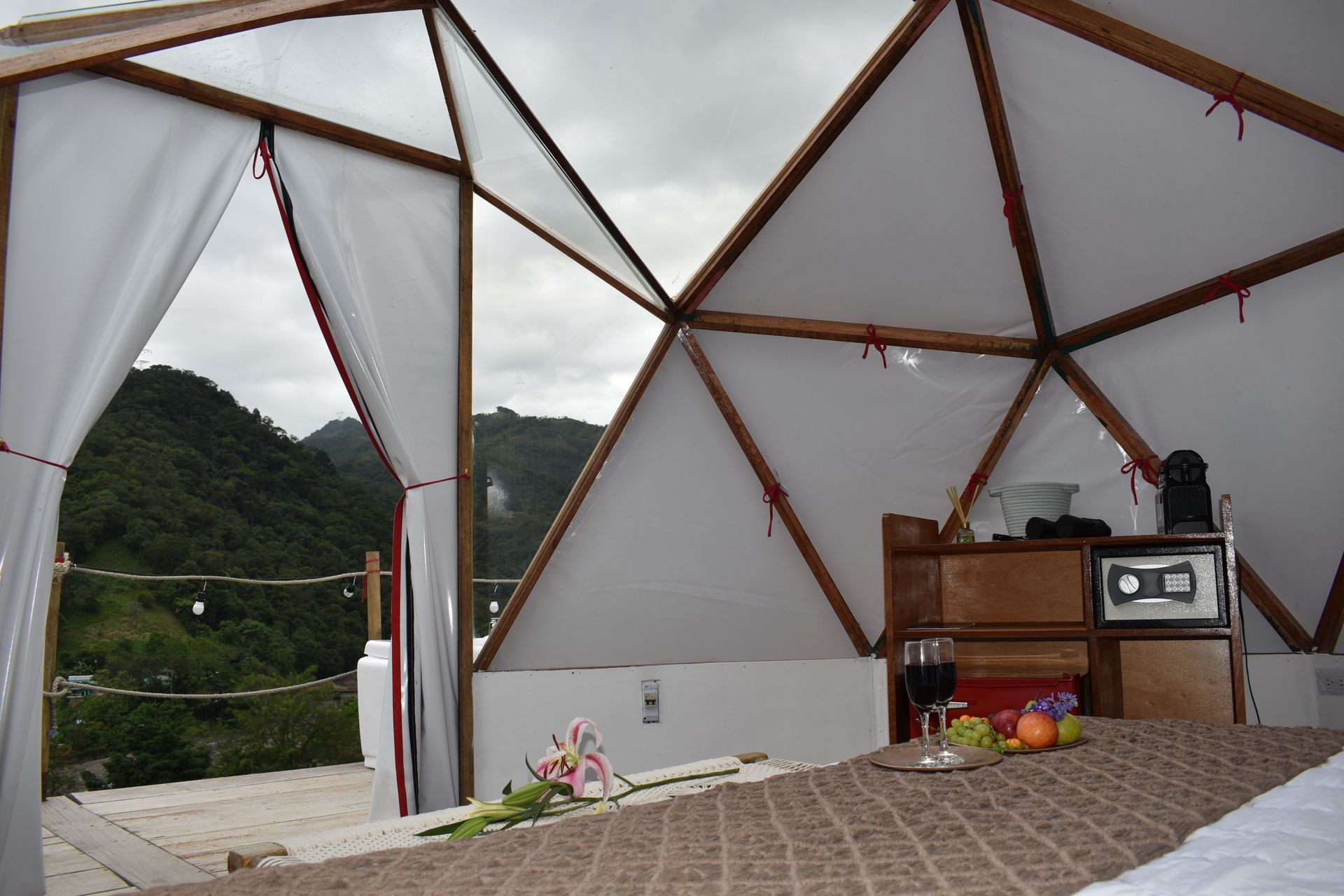 Interior de cúpula geodésica con vista a una verde cordillera, cama con frutas y vino.