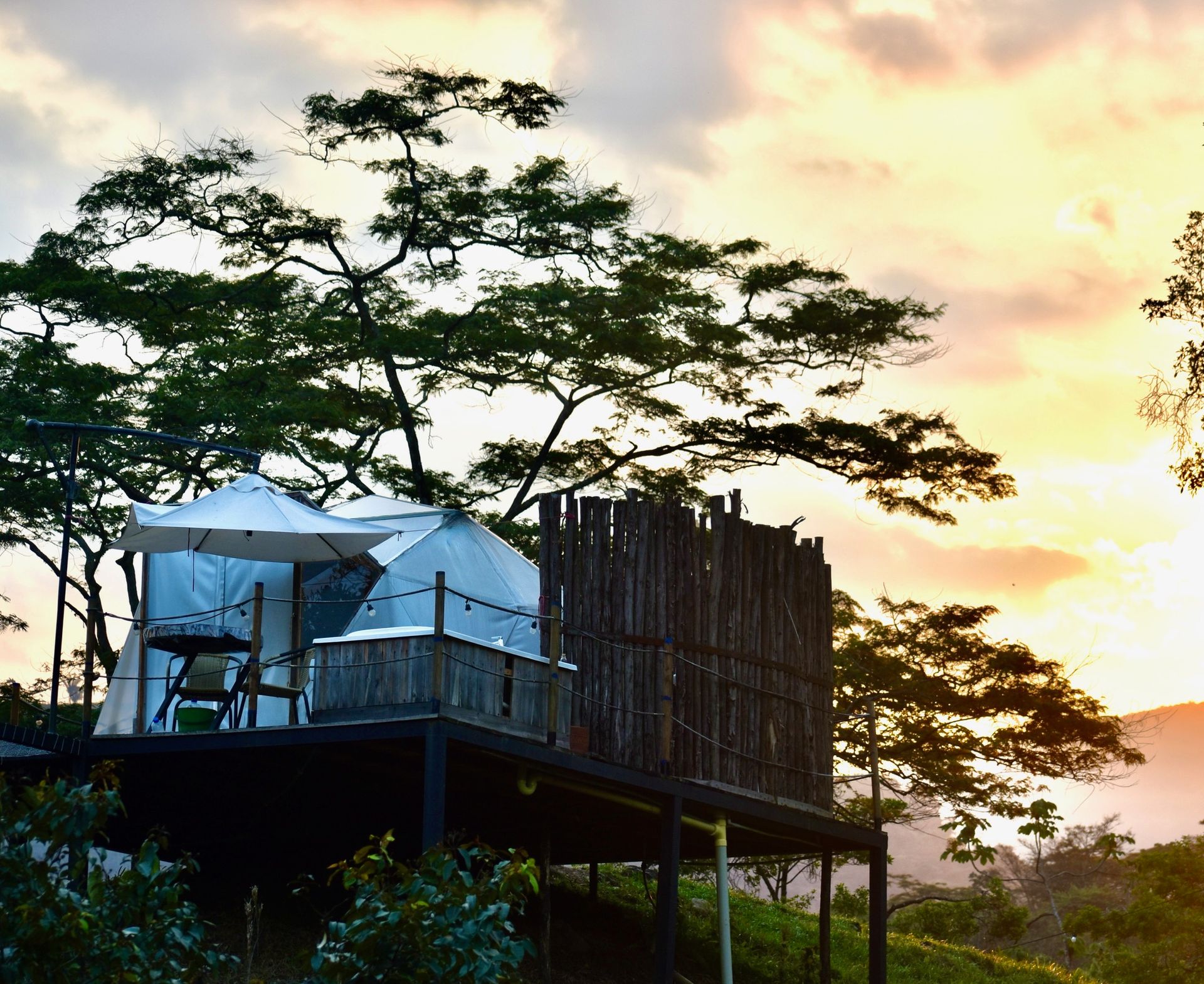Plataforma de cabaña de madera con mesa y sillas, frente a un cielo al atardecer y árboles.