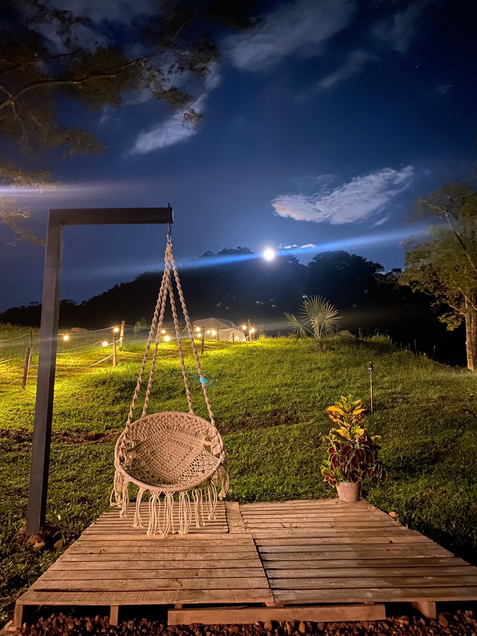 Escena nocturna: columpio, plataforma de madera, luna llena sobre una colina verde, iluminada por luces suaves.