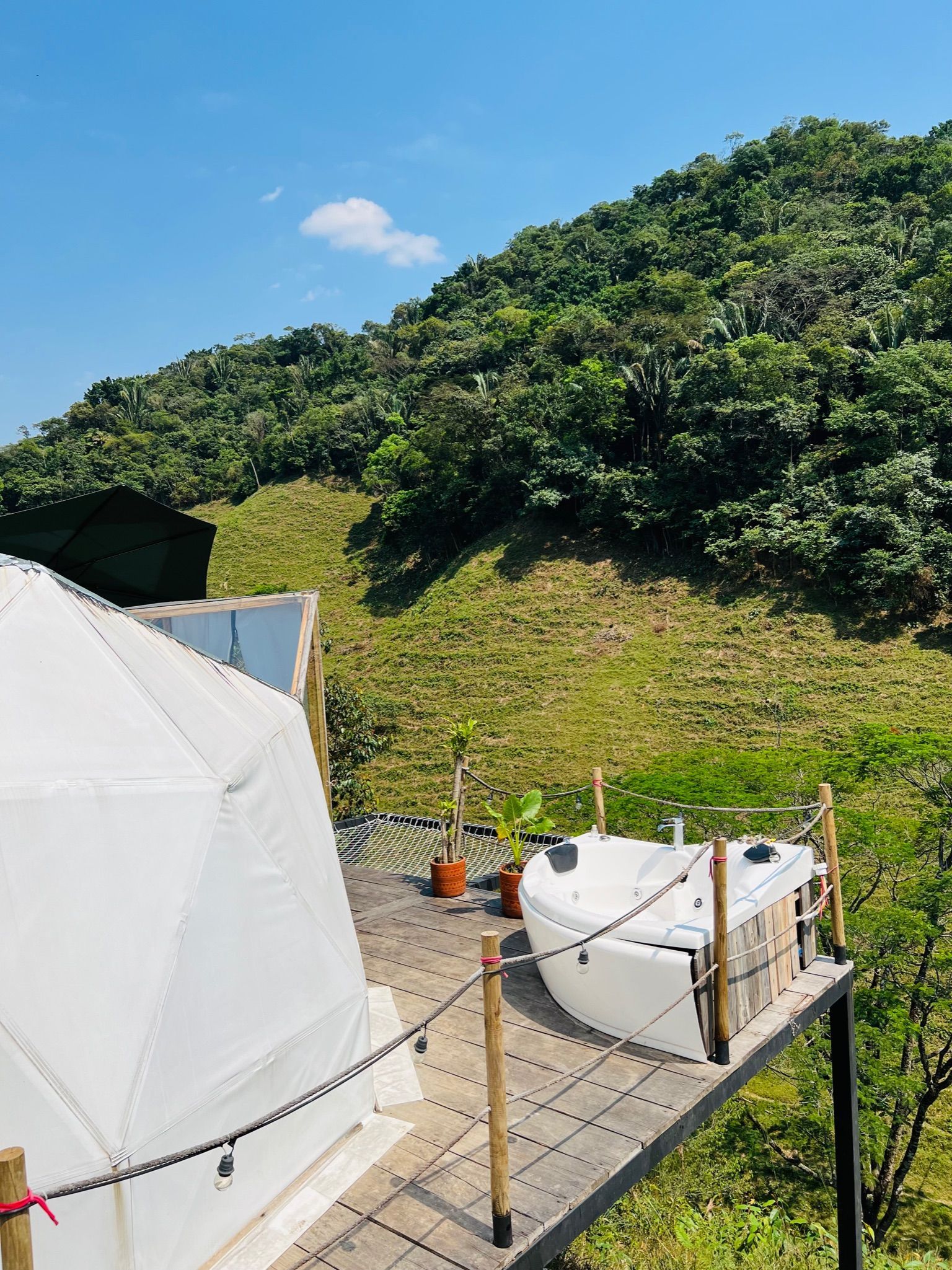Cúpula geodésica blanca con jacuzzi en una terraza con vista a una ladera en un día soleado.