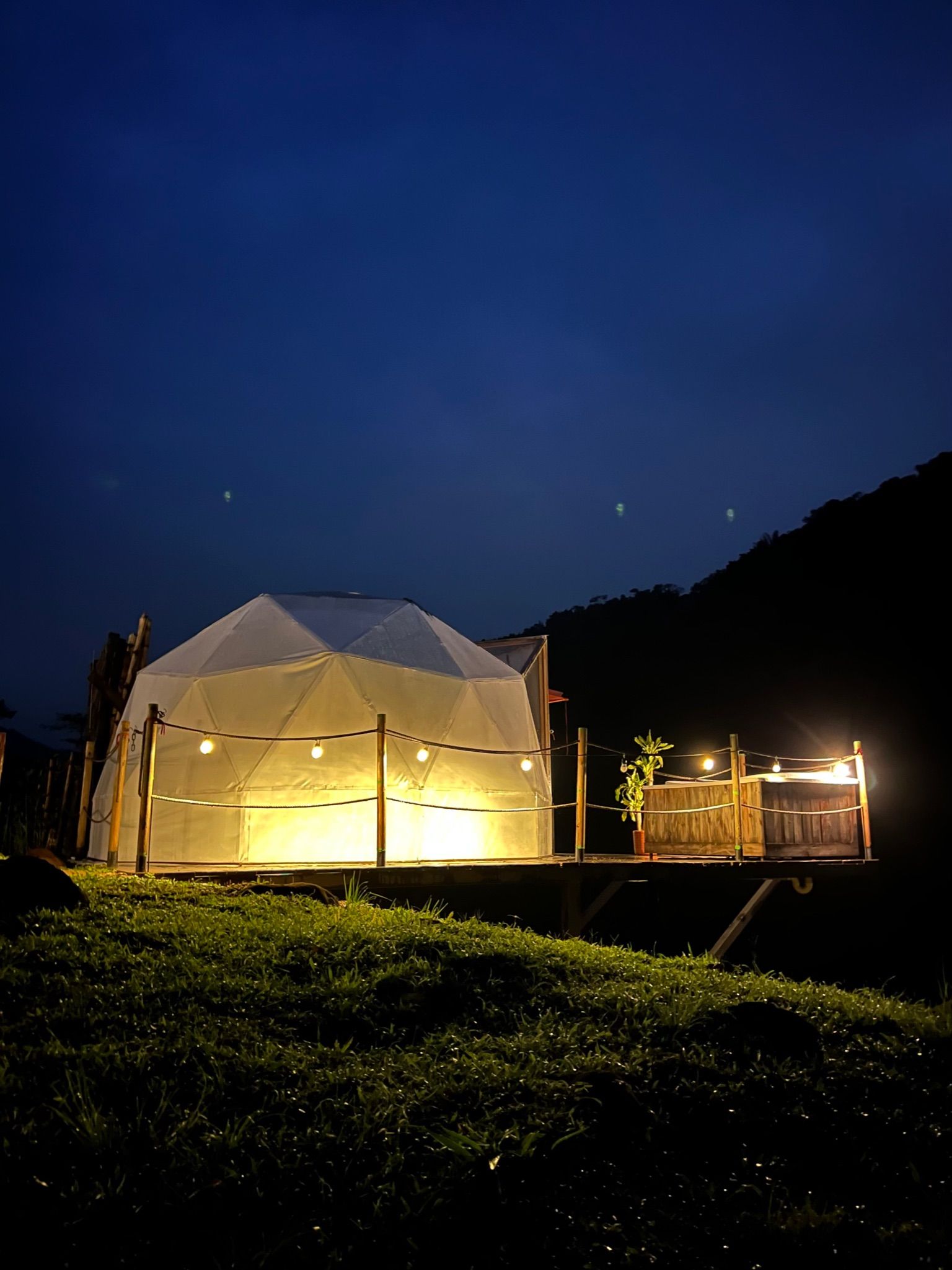 Estructura de cúpula iluminada sobre una plataforma elevada por la noche; cielo oscuro y ladera de fondo.