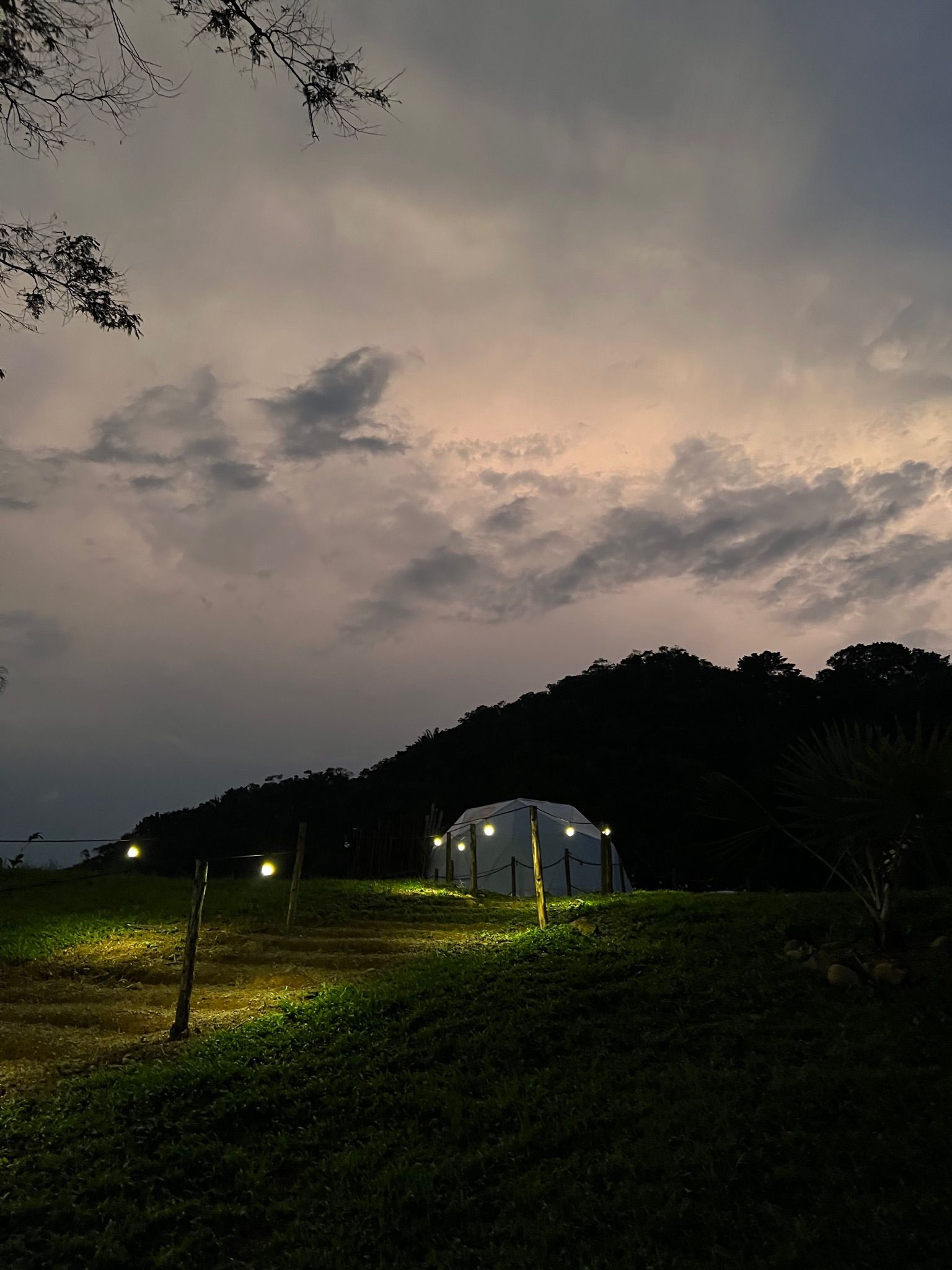Estructura en forma de cúpula iluminada en un campo al anochecer, con una colina recortada y un cielo nublado arriba.