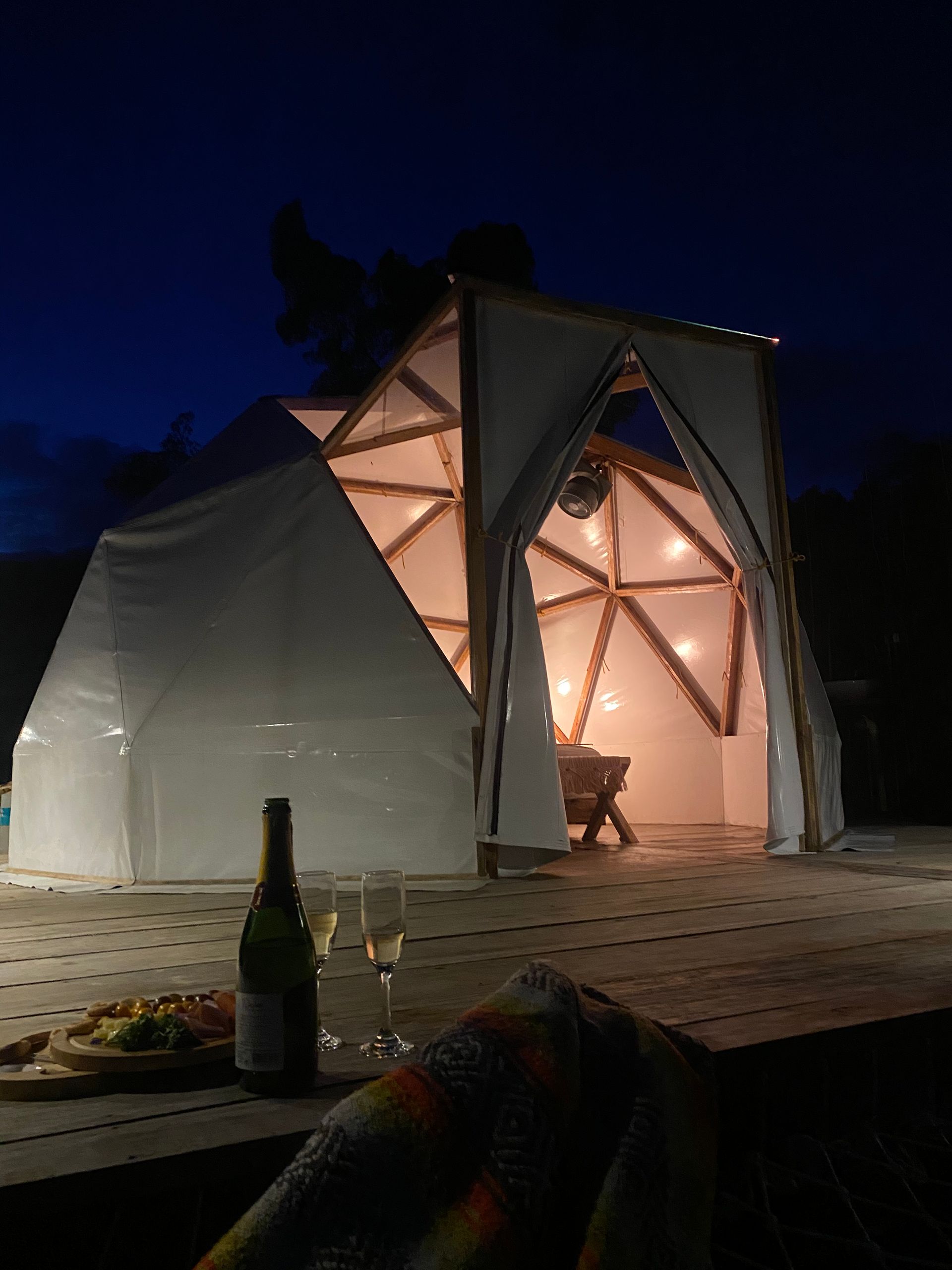 Estructura de cúpula geodésica iluminada por la noche, con comida y bebida en primer plano.