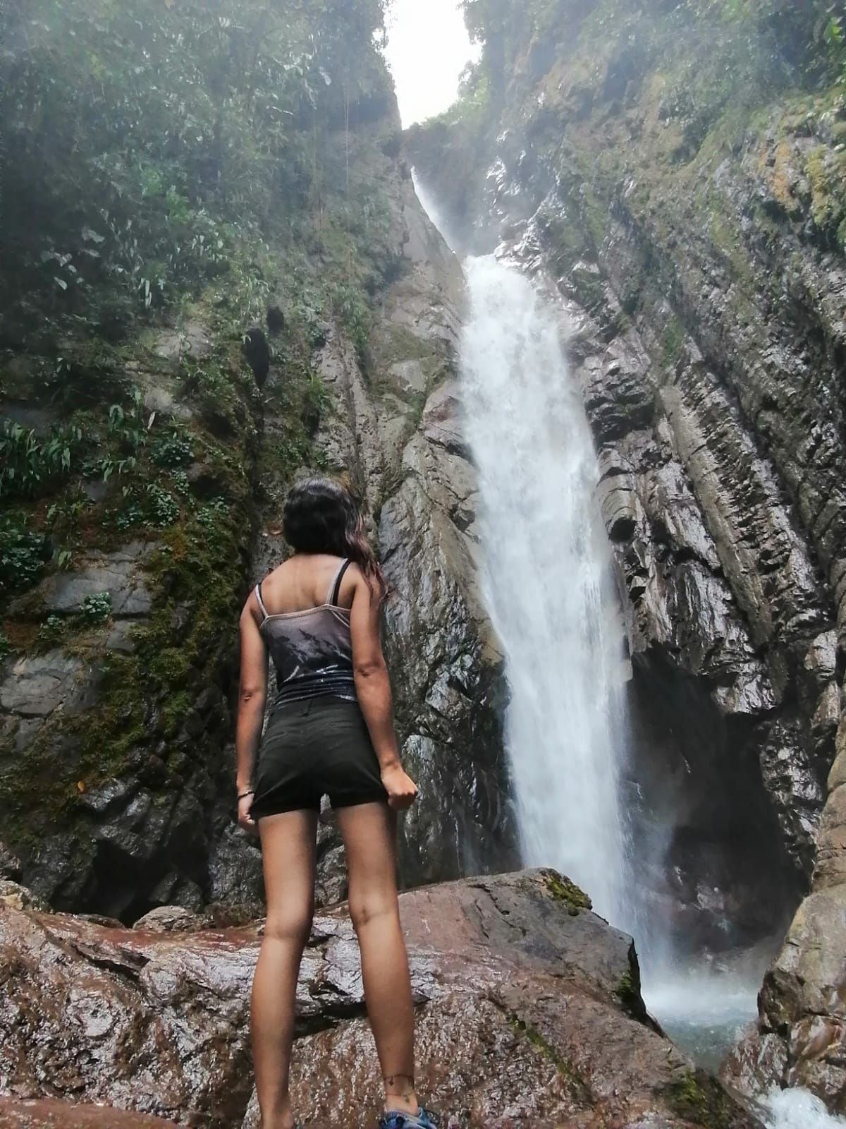 Mujer de pie frente a una cascada en un entorno rocoso y exuberante.