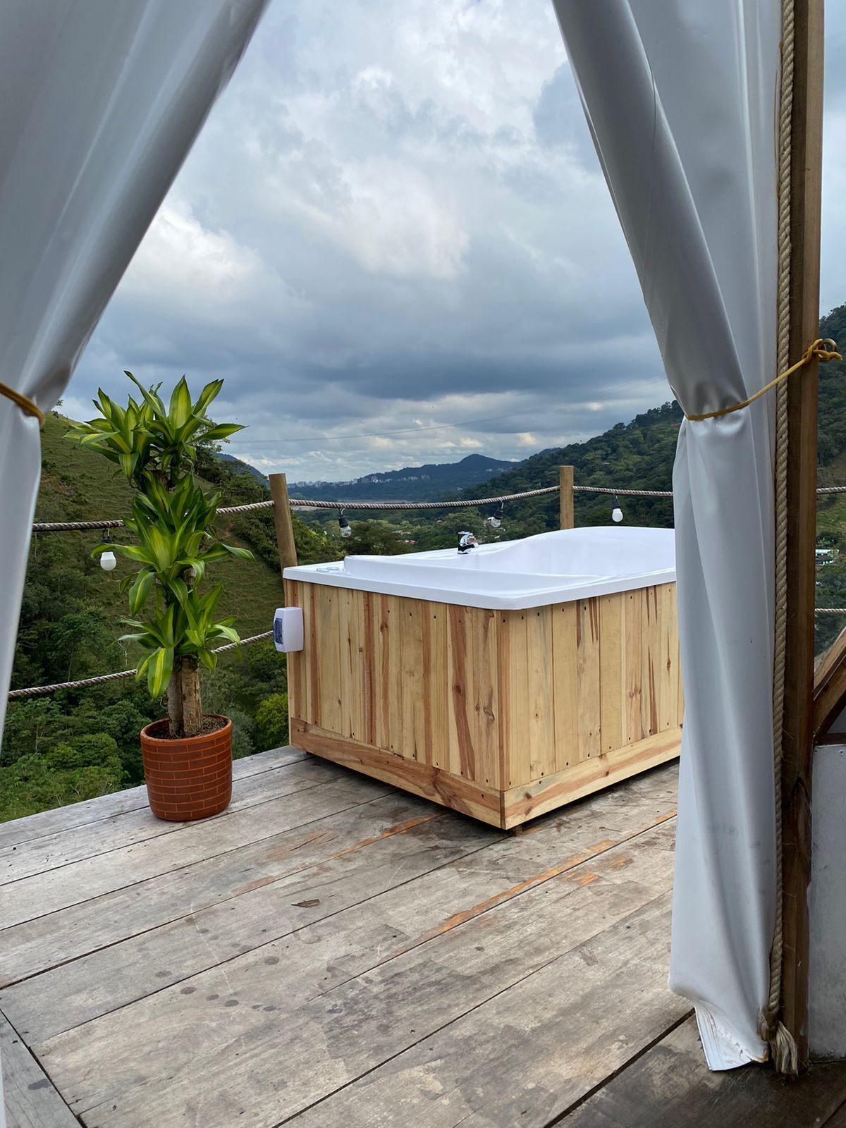 Jacuzzi de madera en una terraza con vistas a la montaña, enmarcado por cortinas blancas y una planta en maceta.