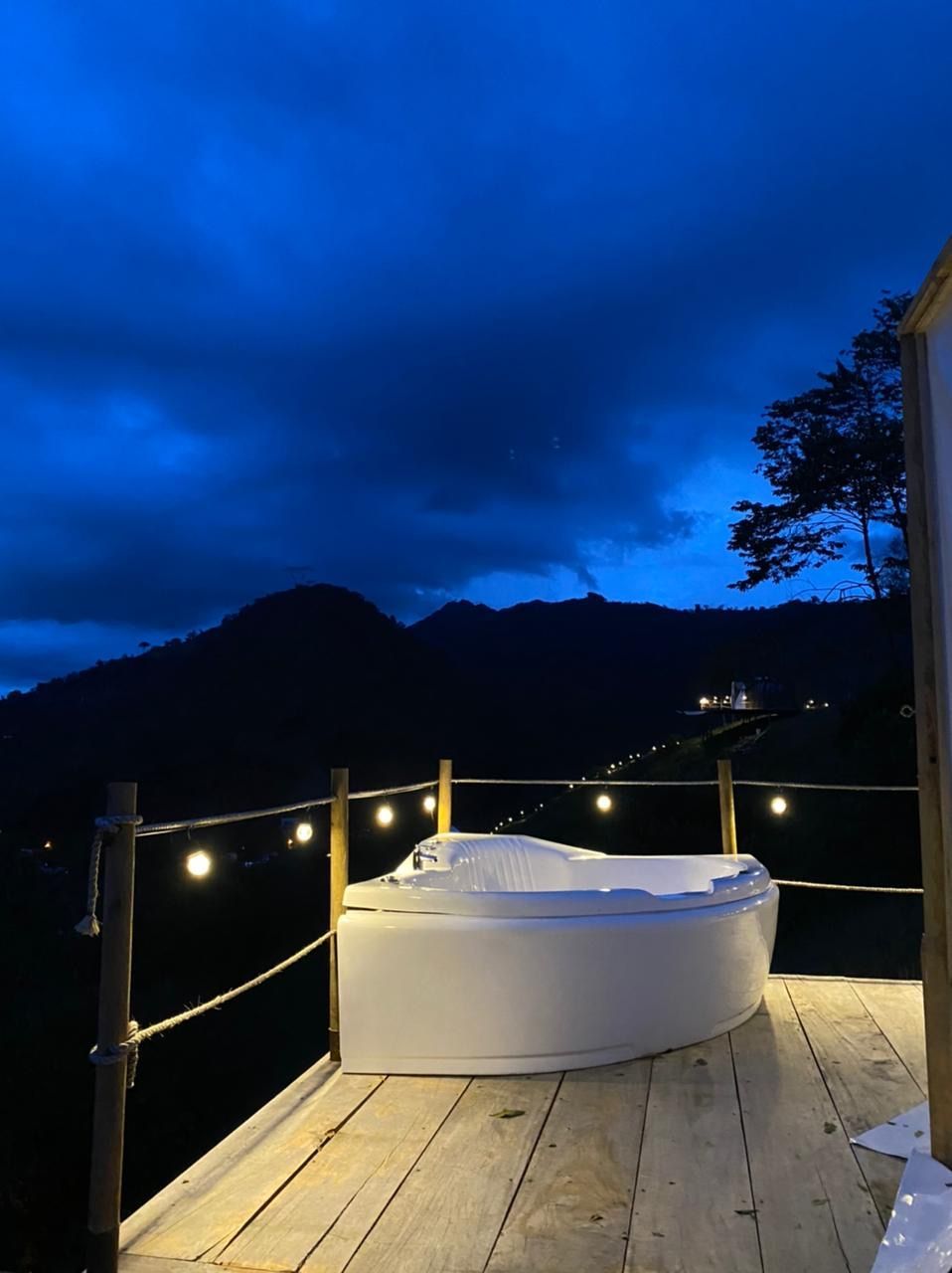 Jacuzzi en terraza de madera, con vistas a las montañas al anochecer. Luces de colores y cielo azul oscuro.