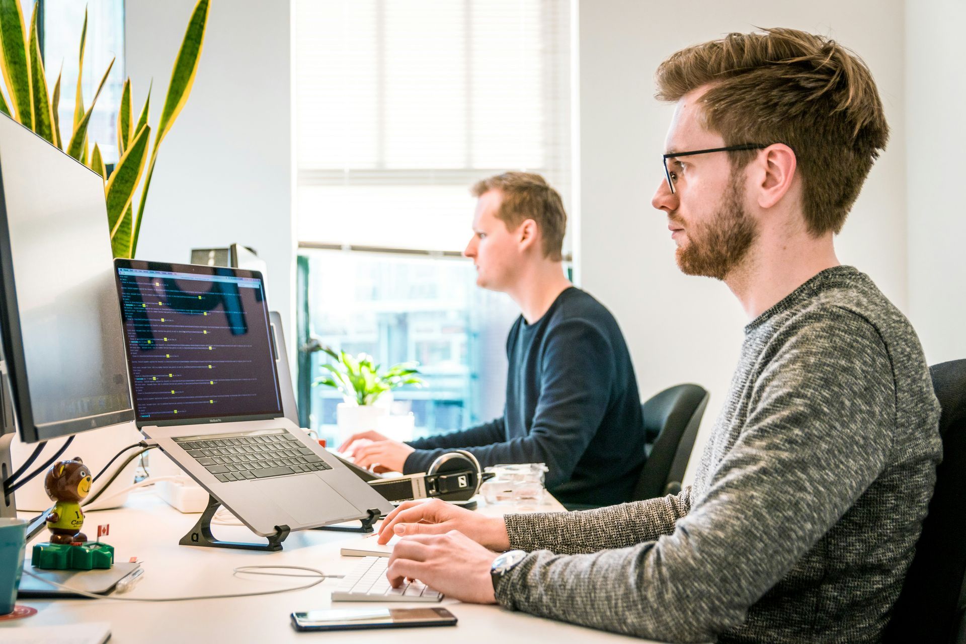 Two men working on computers in an office. One uses a laptop on a stand, the other works on a desktop.