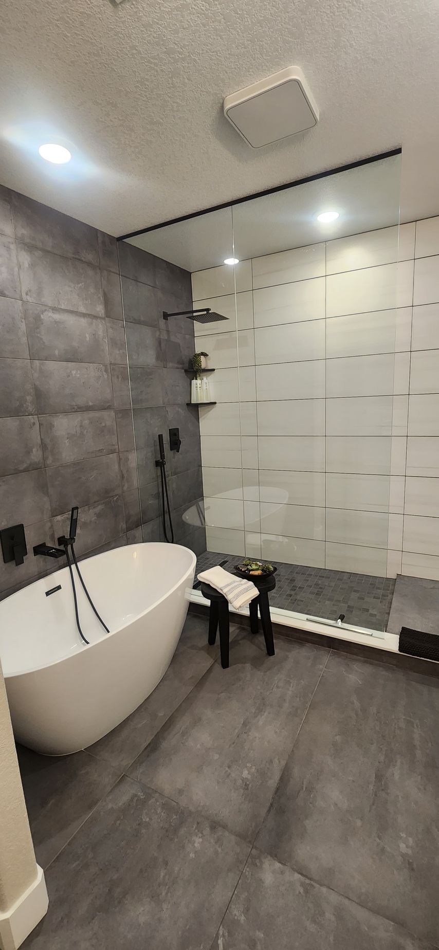 Modern bathroom with a white tub, walk-in shower, and gray tile walls and floor.