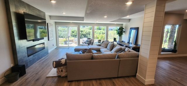 Living room with gray sectional, large windows, fireplace and wood flooring.