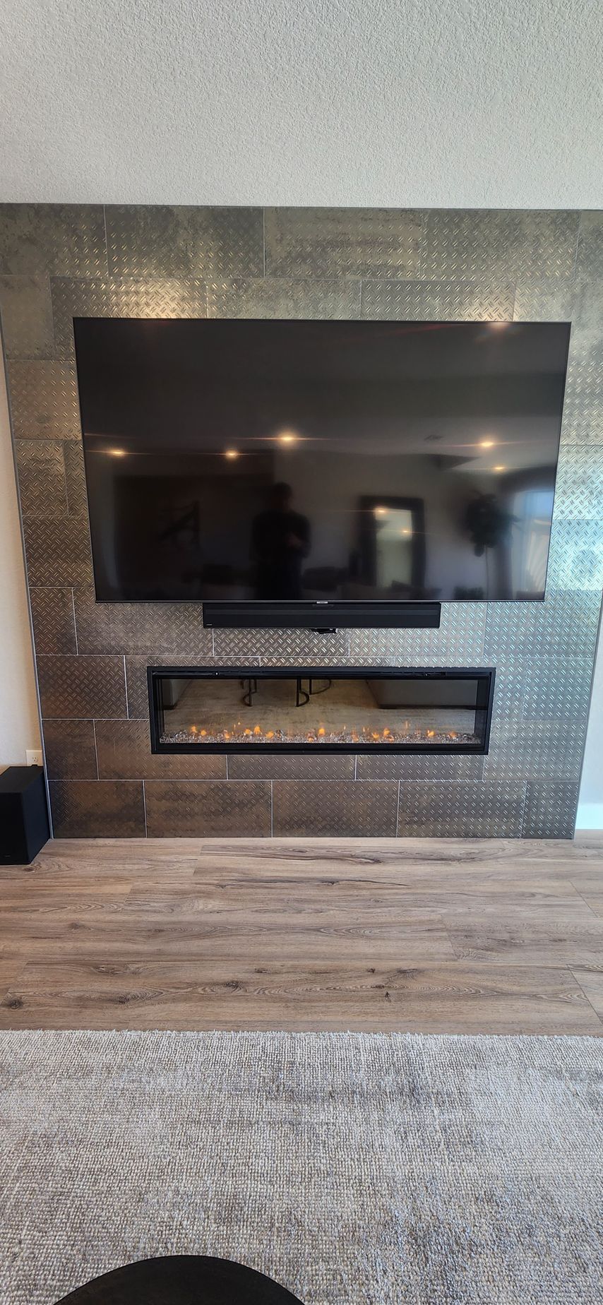 Living room with TV above fireplace, silver tiled wall, dark wood floors, and grey carpet.