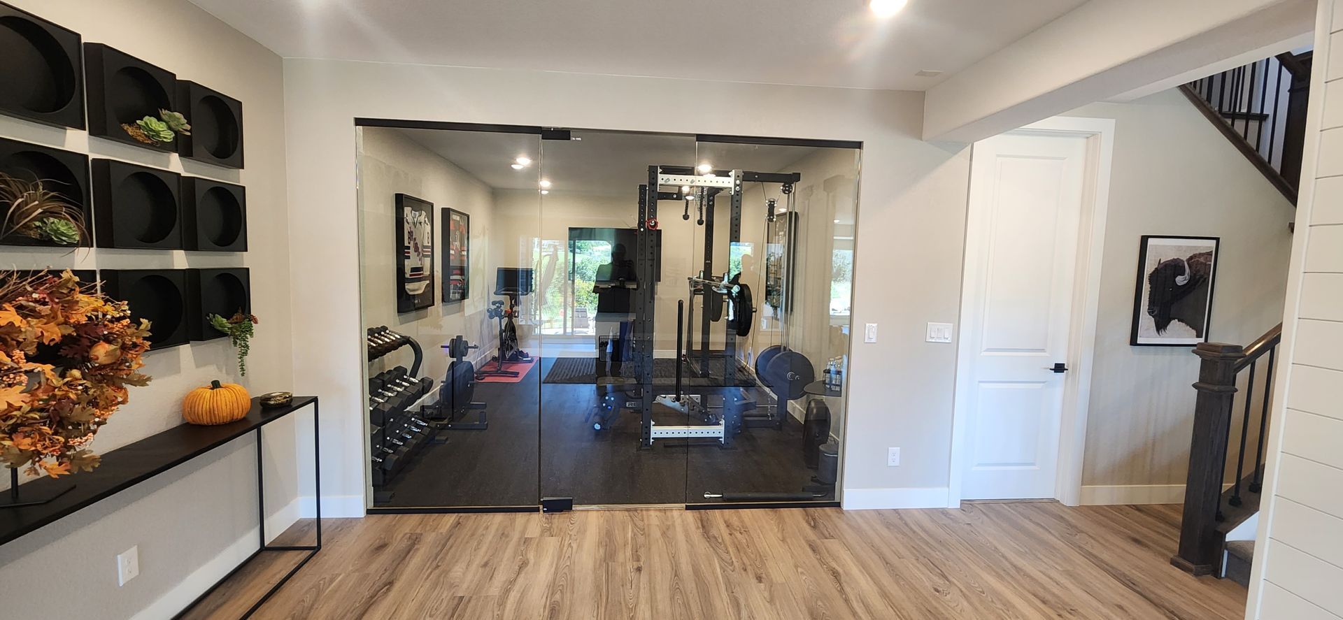 Home gym with glass doors. Weight equipment and a person are visible inside. Wooden floor and stairs.