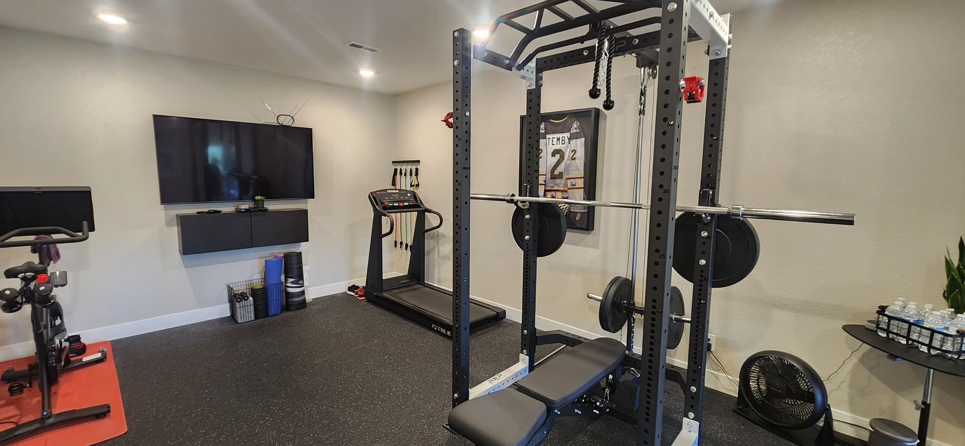 Home gym with weight rack, treadmill, and stationary bike. Black rubber flooring, TV mounted on the wall.