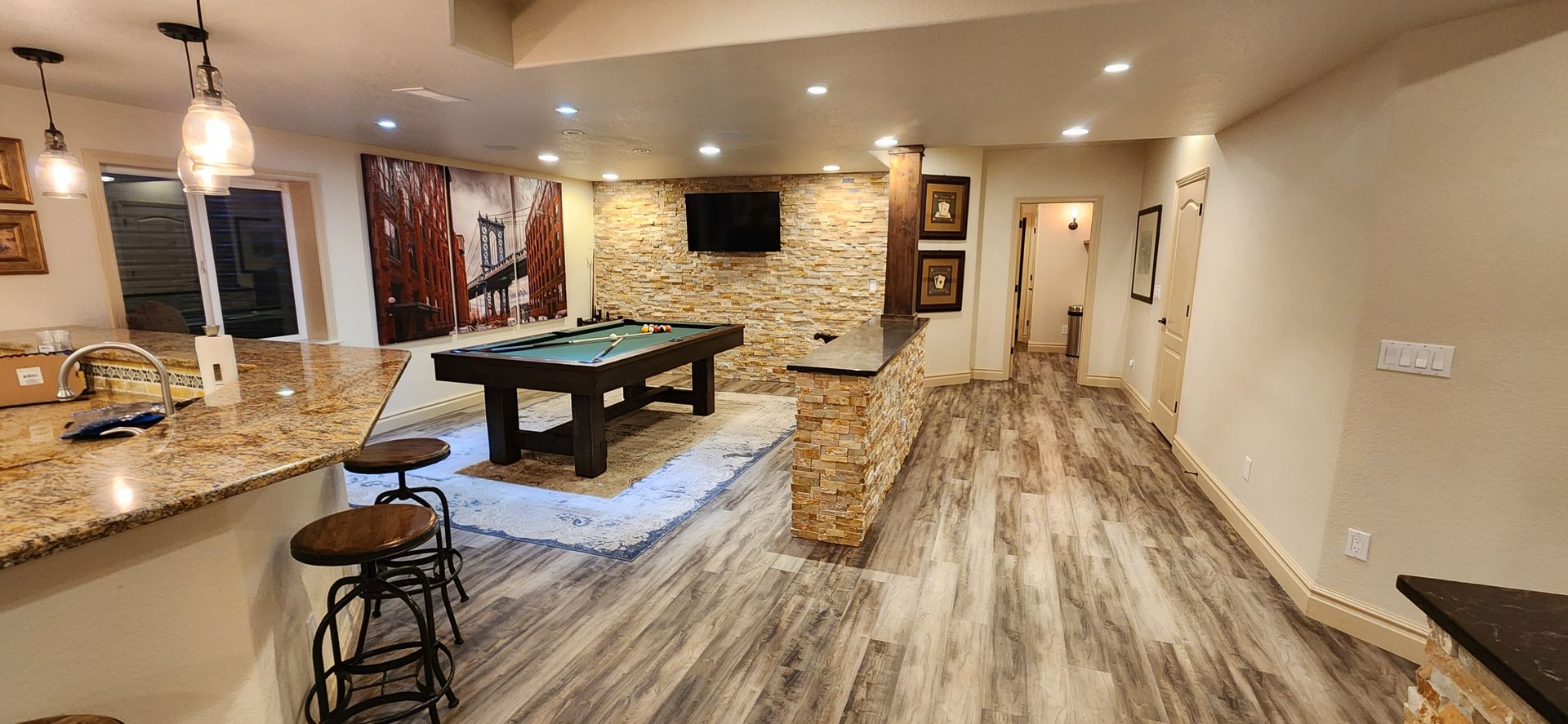 A finished basement with a pool table, bar, stone wall, and wood-look flooring.