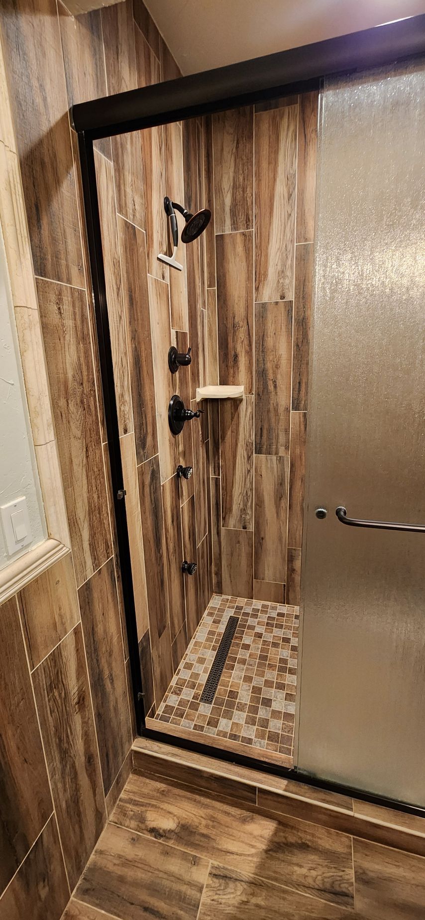 Shower with dark fixtures, stone tile walls and flooring, and a frosted glass door.