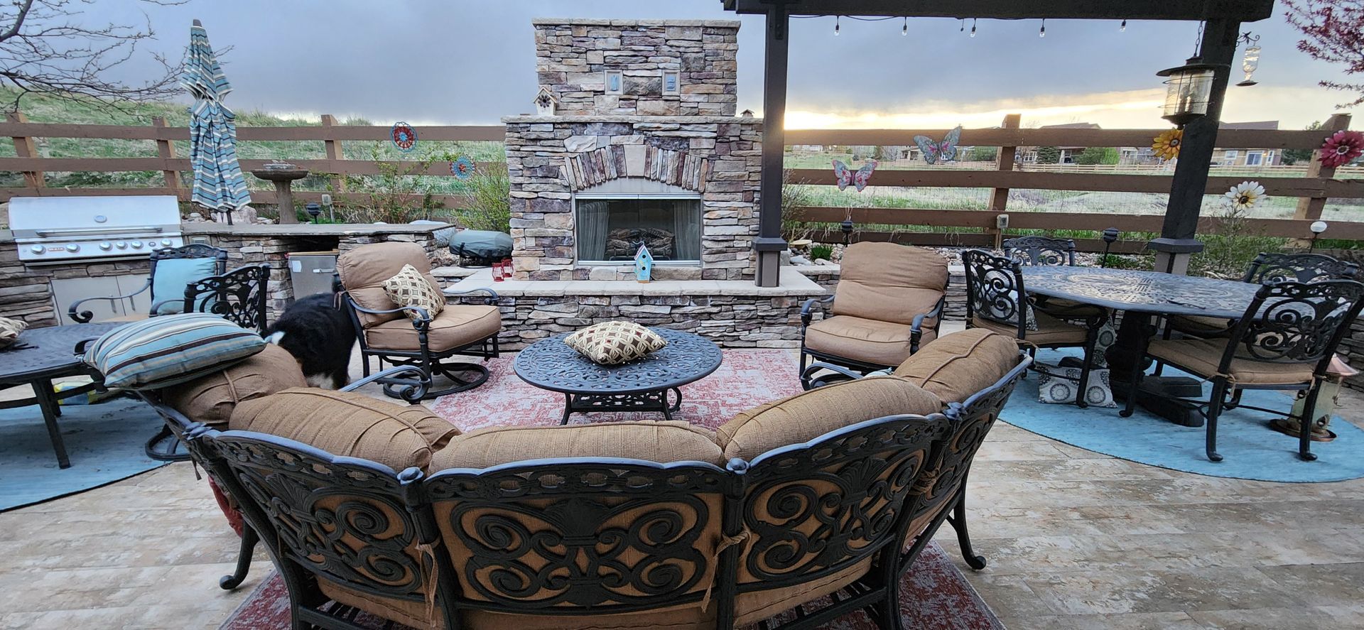 Outdoor patio with stone fireplace, seating area, and dining table. Fenced background and cloudy sky.