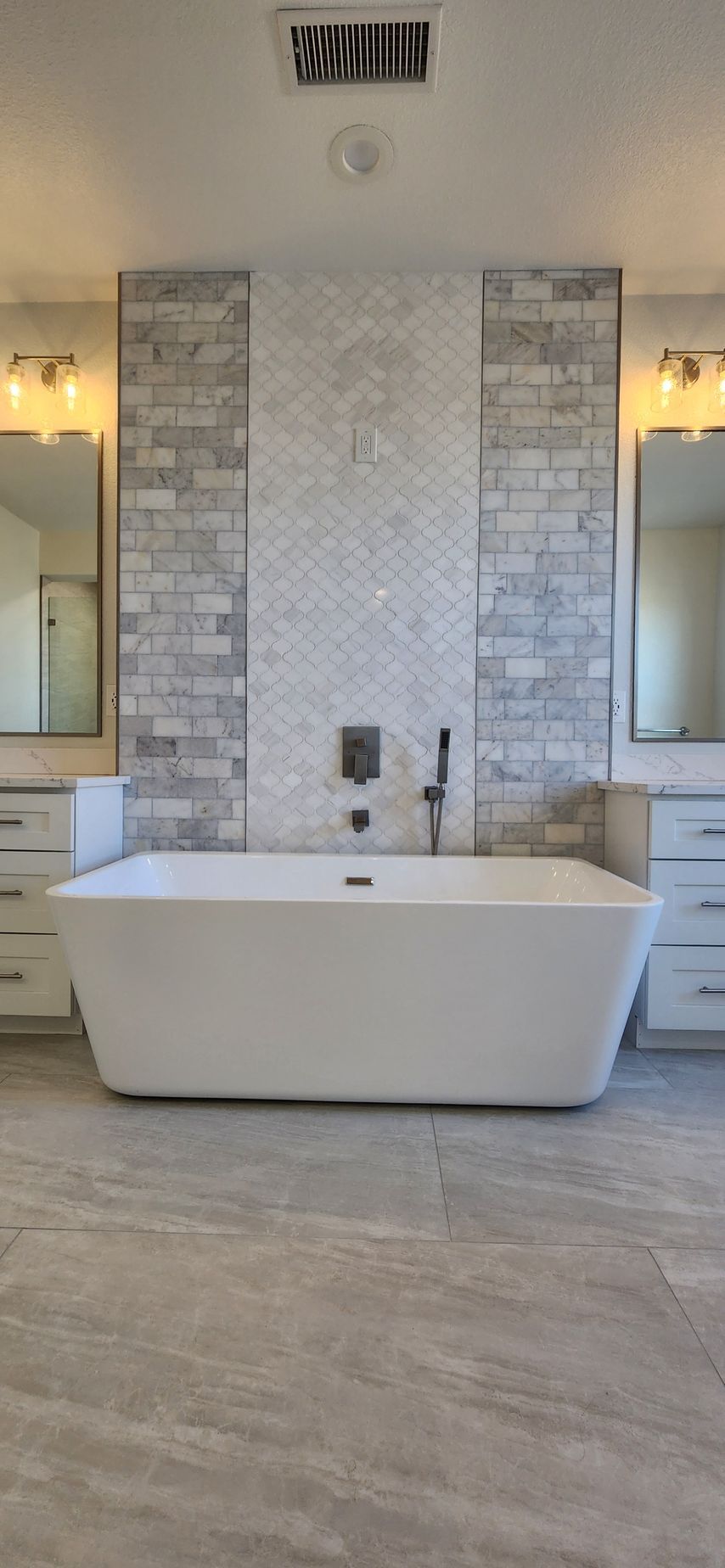 White bathtub centered in a modern bathroom with grey tile floors and a patterned backsplash.