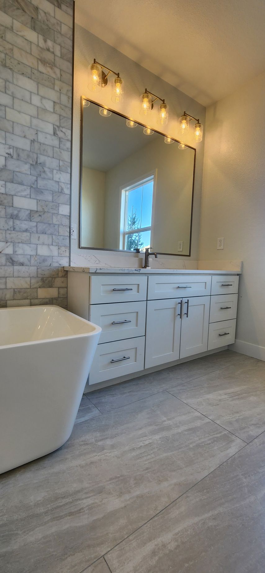 Bathroom with a white vanity, large mirror, and brick-style wall. Gray flooring and a bathtub are visible.