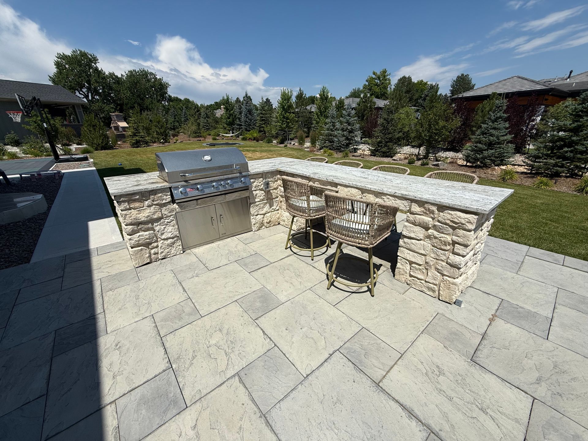 Outdoor kitchen with stone counters, grill, bar stools, on a stone patio with greenery and blue sky.