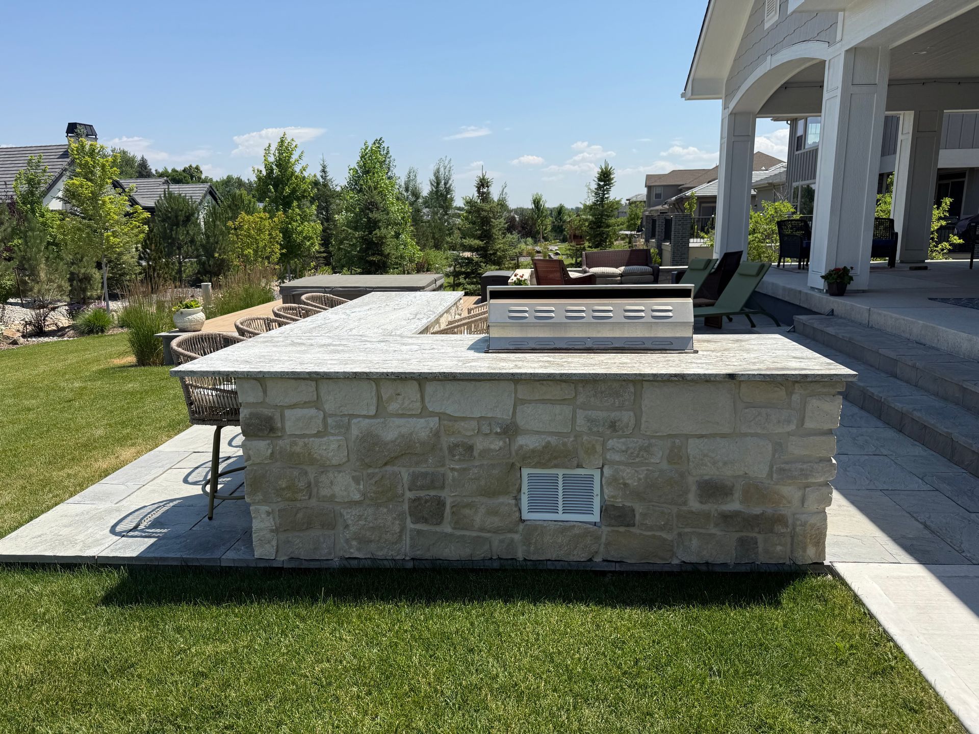 Outdoor stone BBQ island on patio with stainless steel grill. Green grass and trees in background.