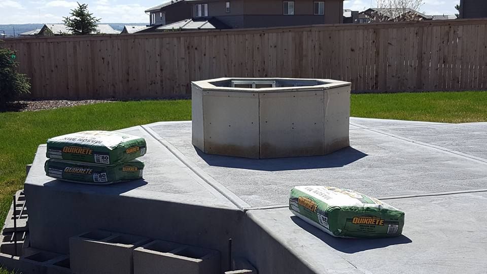 Concrete fire pit on a concrete patio with bags of concrete mix. Wooden fence and grass in background.