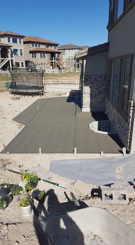 Newly poured concrete patio next to a house under construction; wheelbarrow, plants in pots, and neighborhood in the background.