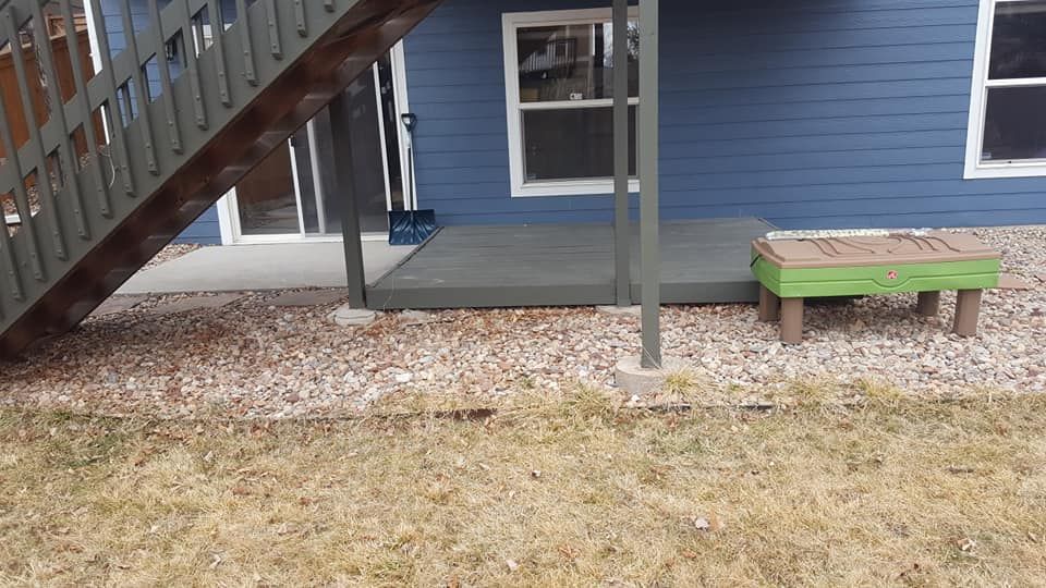 Backyard view: Blue house, wooden deck, stairs, rock bed, and small green and brown table.