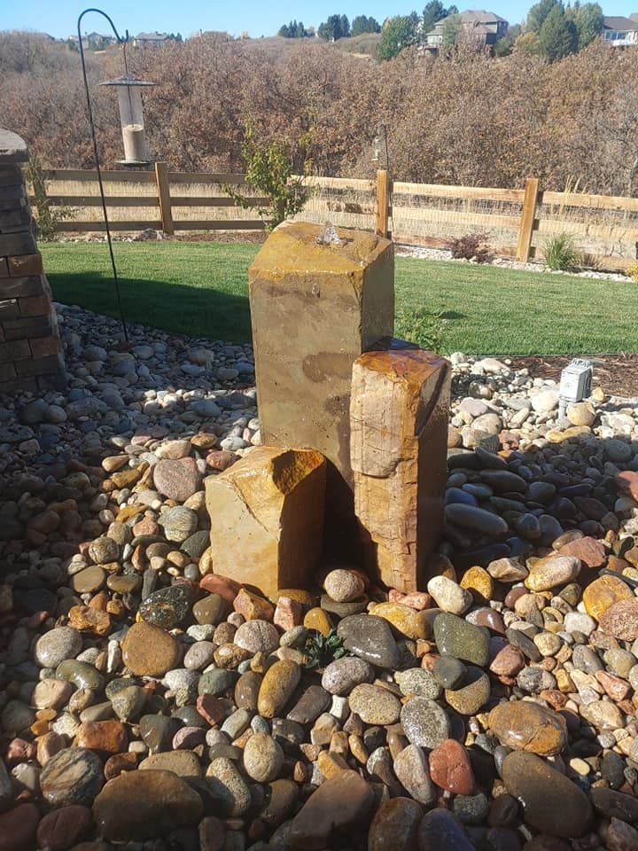 Fountain with three stone pillars among wet river rocks, in a yard with a grassy area and fence in background.