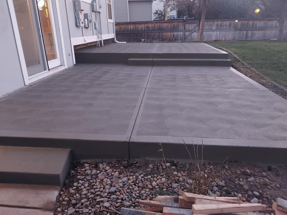 Newly poured gray concrete patio with steps leading to the backyard, next to a house with an open doorway.