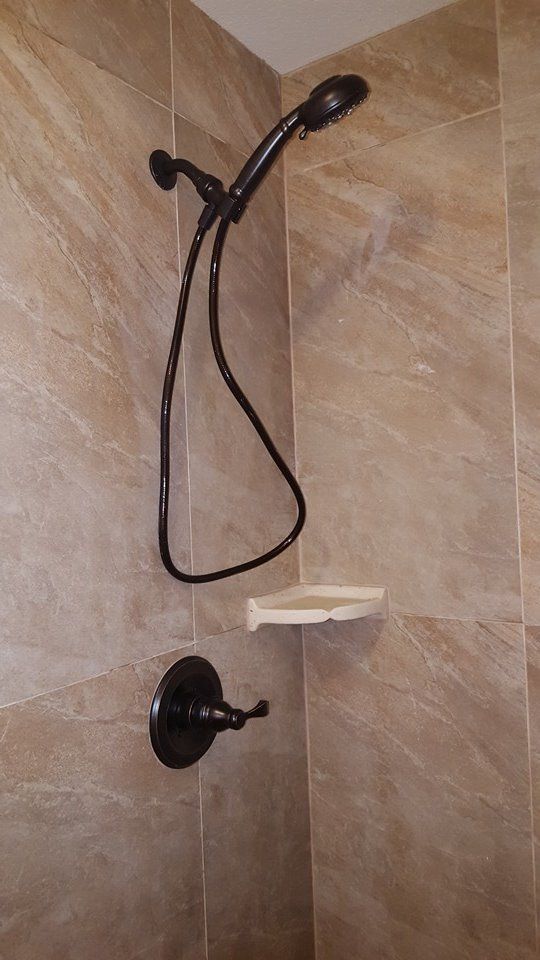 Dark-colored shower fixtures on beige-tiled wall. Includes handheld showerhead, shelf, and faucet handle.