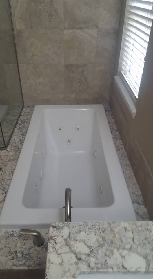 A built-in white jacuzzi tub with silver fixtures, surrounded by beige tile and granite countertops, near a window.