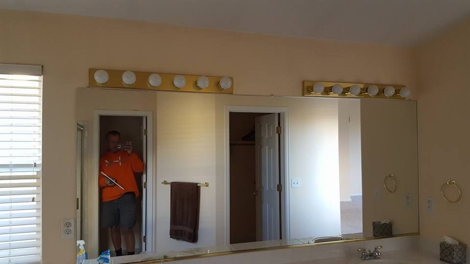 A man in an orange shirt holds an item near a bathroom mirror; lit by two fixtures.