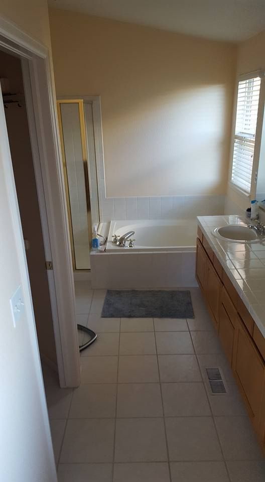 Bathroom with a bathtub, vanity, and closed door. Beige walls, tile floor, and a window with blinds.