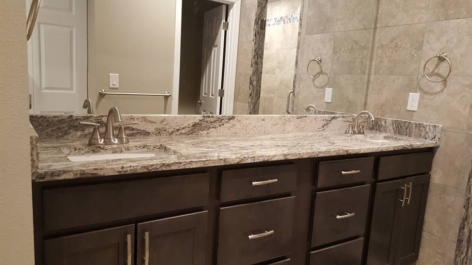 Bathroom with dark cabinets, granite countertop, and dual sinks with large mirror and light-colored tiled walls.