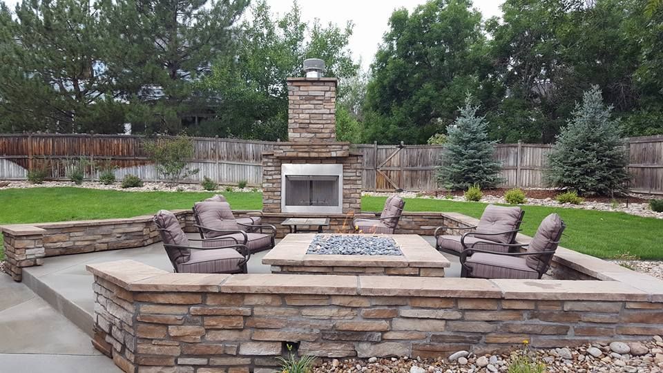 Outdoor stone fireplace with seating around a fire pit on a patio, surrounded by a lawn and trees.