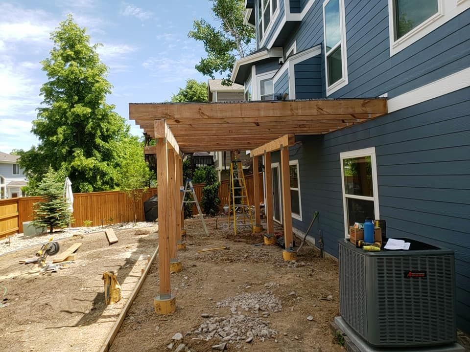 Pergola construction attached to a blue-sided building with windows, set on a dirt yard.