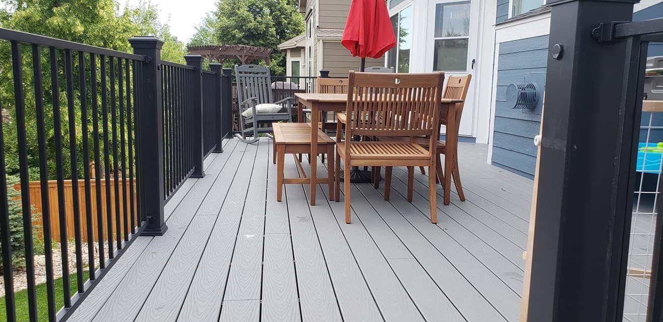 A deck with gray planks and black railing, has wooden furniture with a red umbrella.
