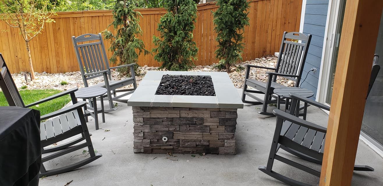 Outdoor patio with a stone fire pit surrounded by gray rocking chairs. A wooden fence is in the background.