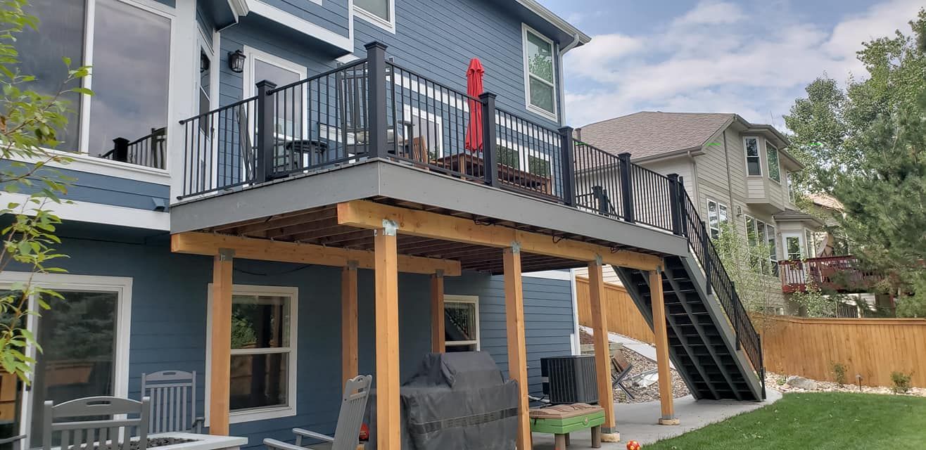 Blue house with a deck and black railing. Staircase leads down to the yard with grass and a wooden fence.