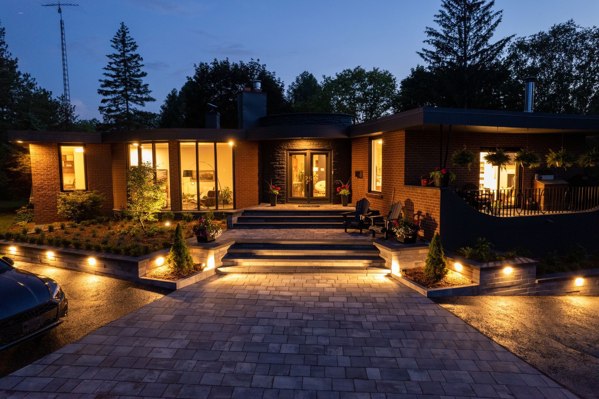 Modern house exterior lit at night. Stone walkway leads to the entrance. Windows glow with interior light.