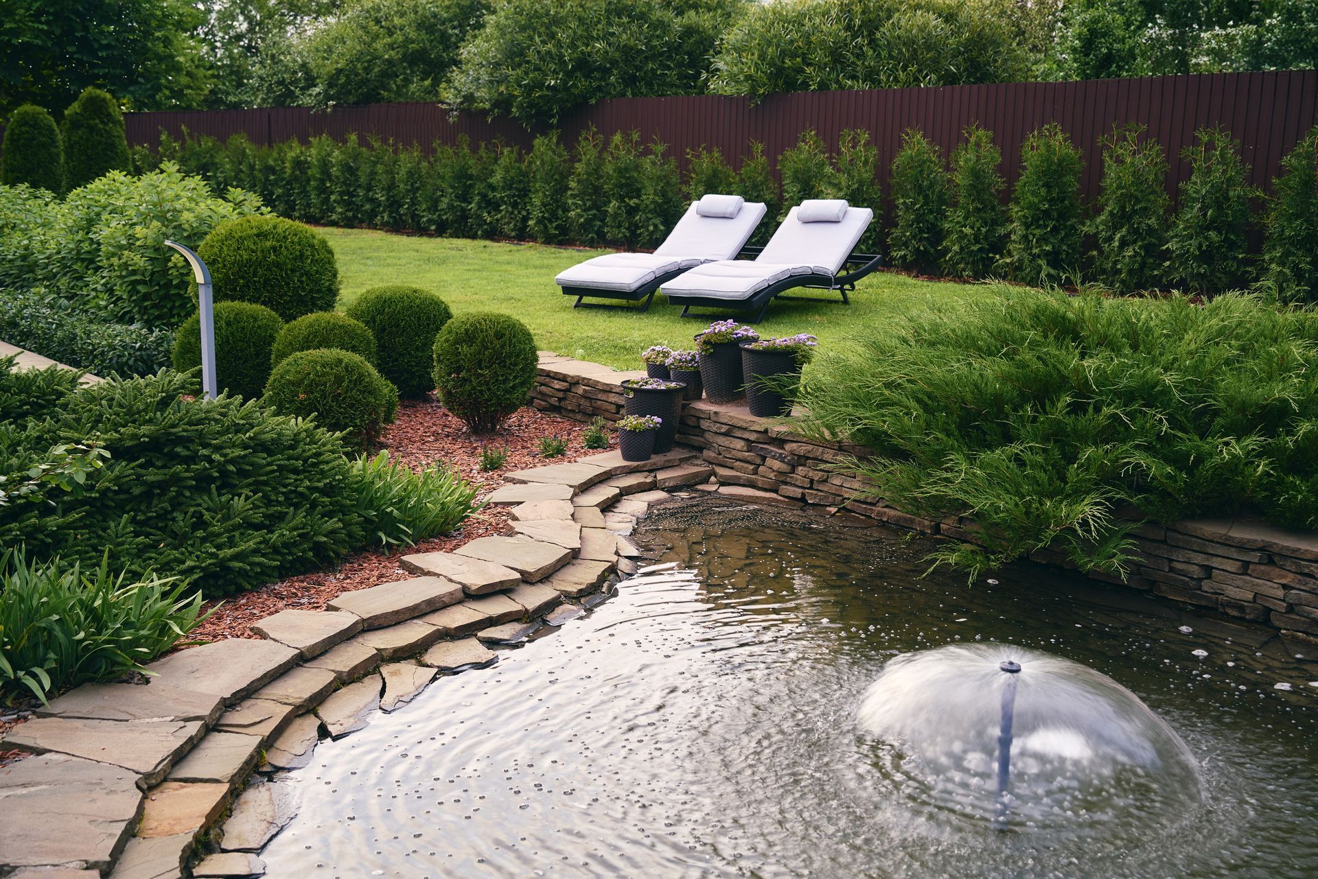Lush garden with a pond and fountain. Two lounge chairs sit on the grass.